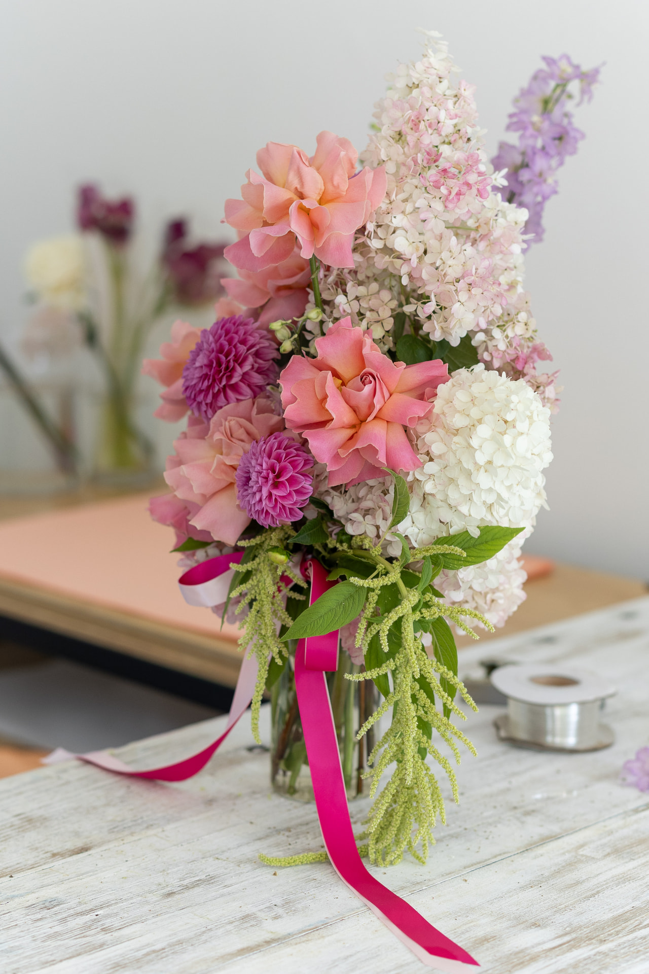 Tall vase arrangement of peach roses, dahlias, hydrangeas and delphiniums with trailing ribbons on the workbench