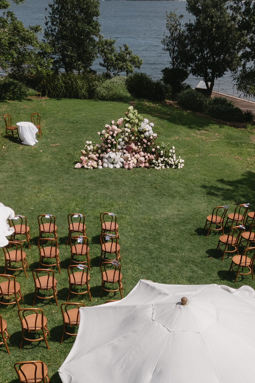 Aerial view of a wedding ceremony on a lawn by the water, with a lush floral meadow of hydrangeas, roses and amaranthus surrounded by bentwood chairs
