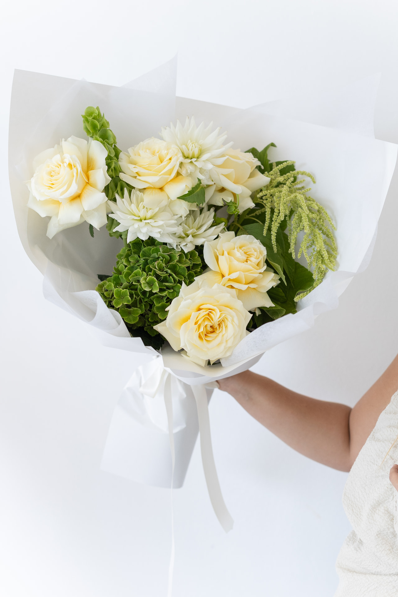 Hand holding a white and yellow bouquet of roses, dahlias and hydrangea wrapped in white paper