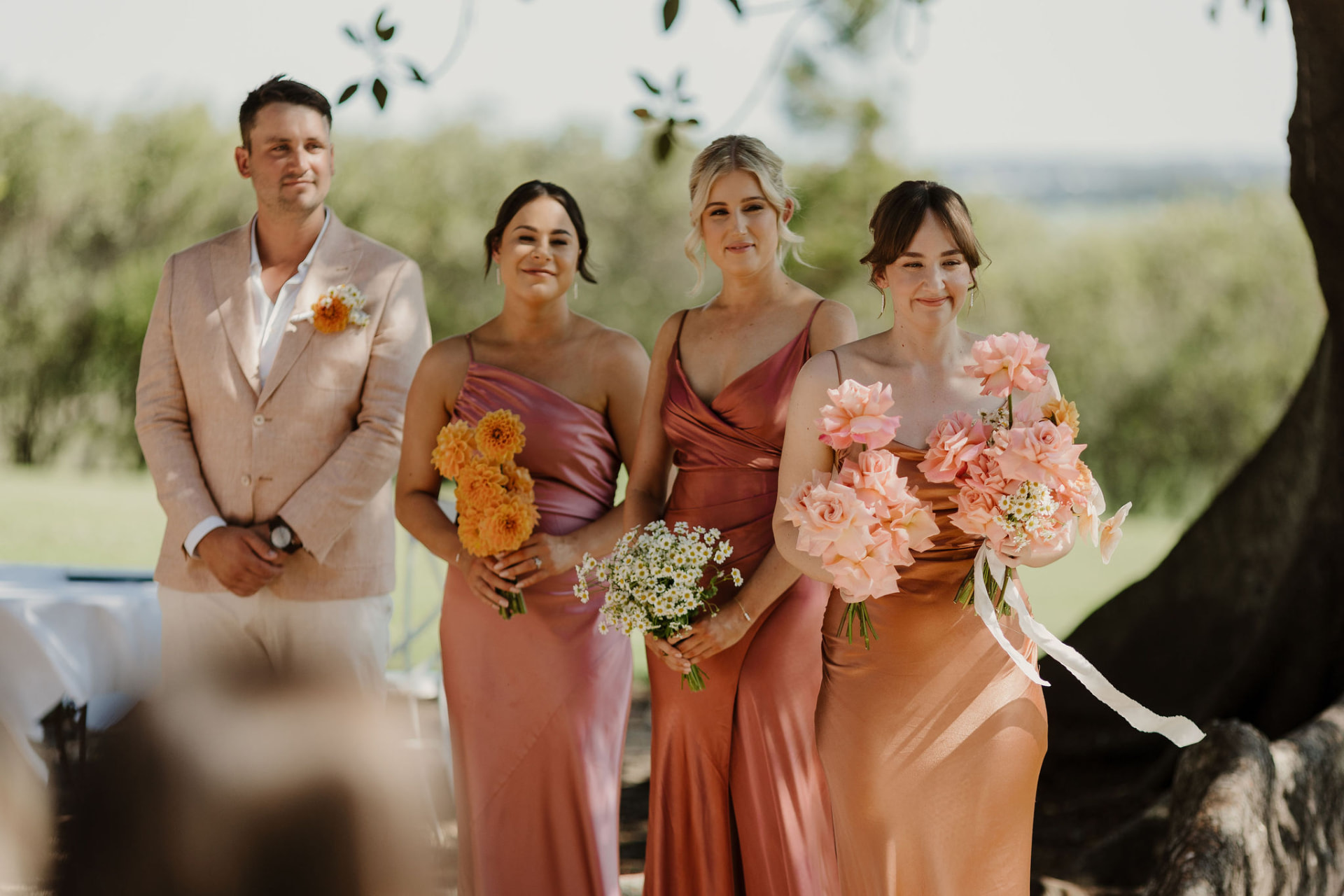 The bridal party in rust and rose silk dresses holding bouquets of peach roses, dahlias and daisies