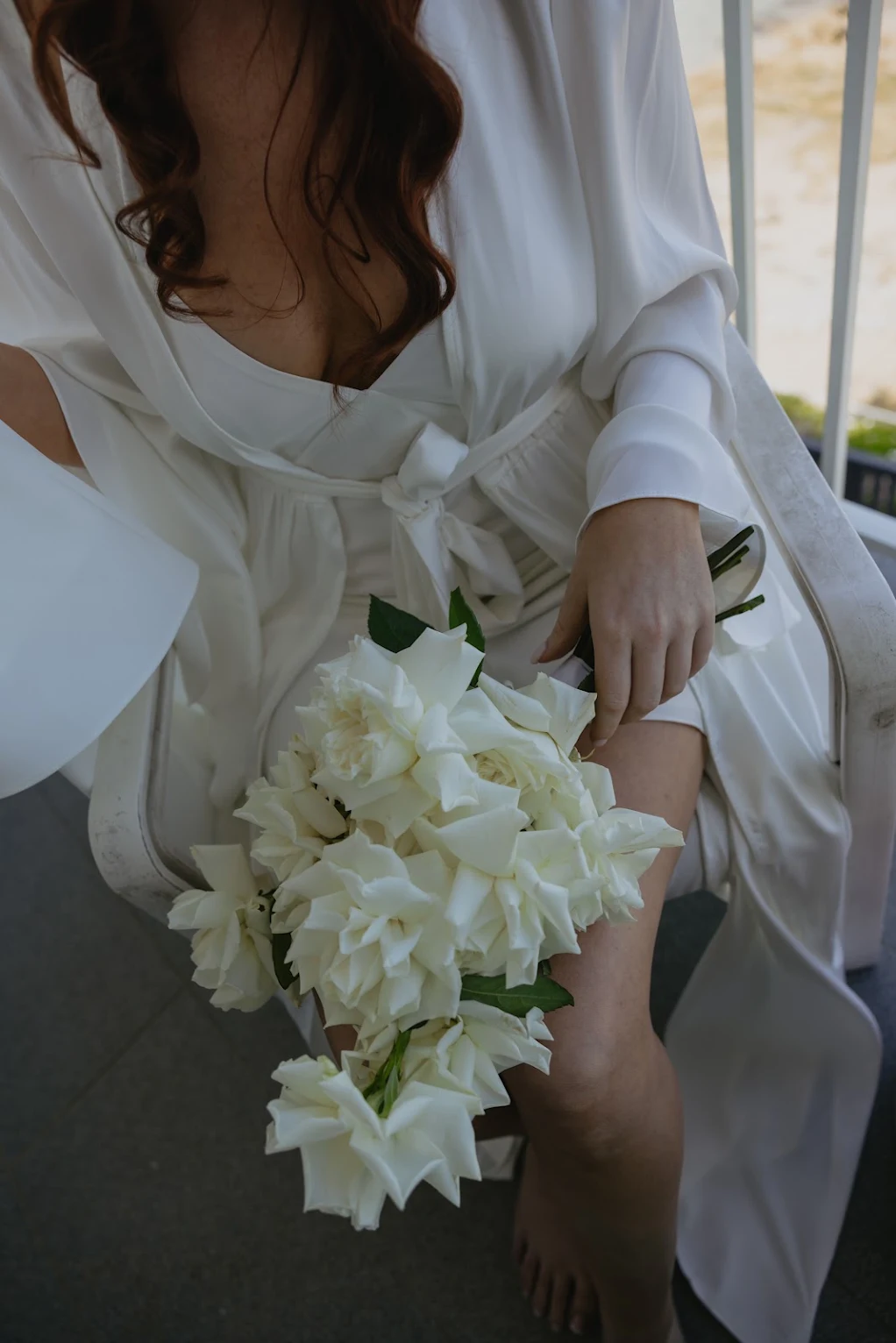 Bride in a white robe holding a loose bouquet of white roses on her lap