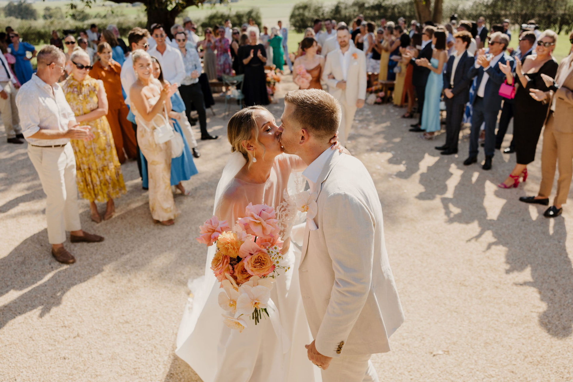 Zoe and Adam kissing after their ceremony, Zoe holding a bouquet of peach roses, orchids and daisies