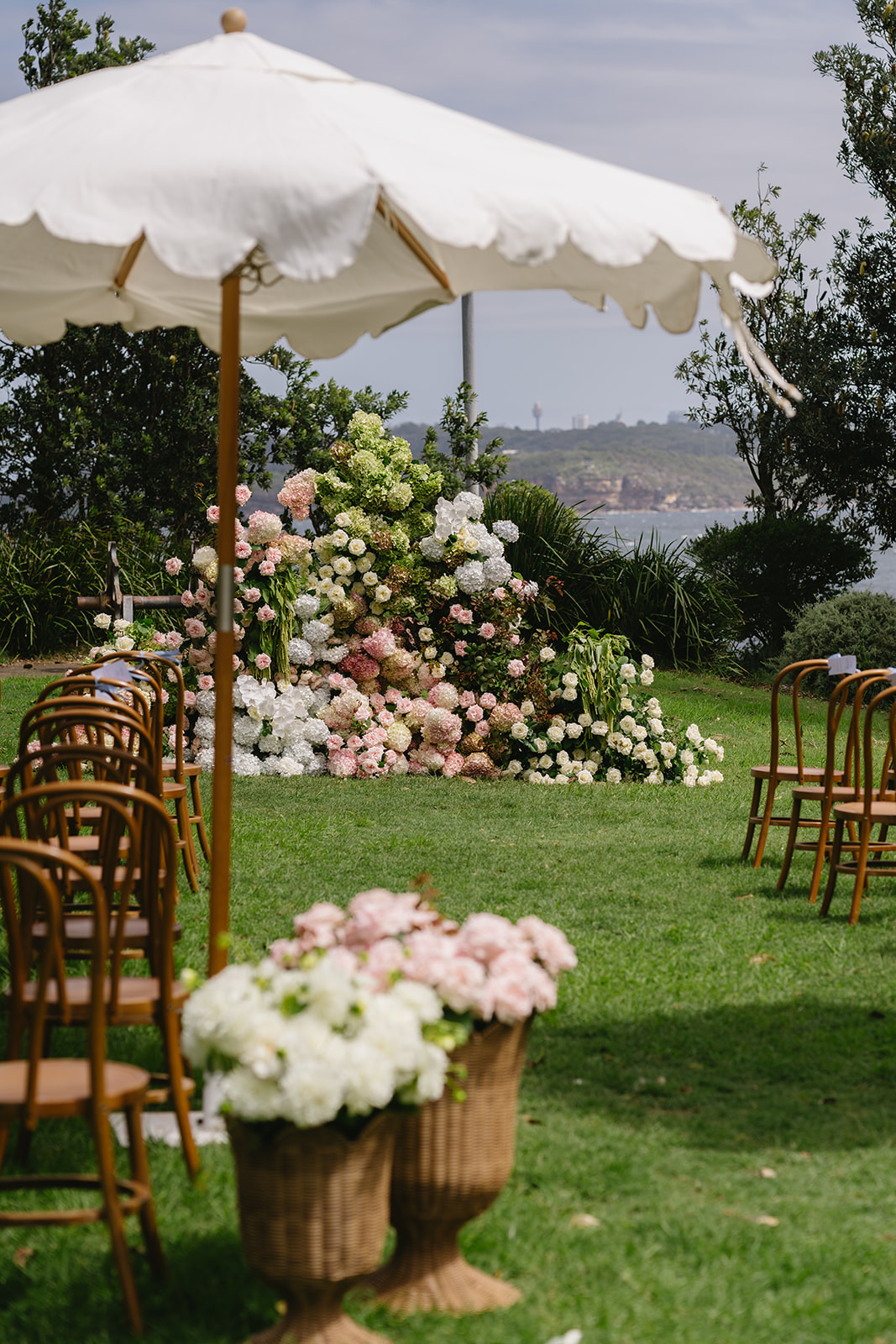 Bentwood chairs facing a floral ceremony meadow beneath scalloped market umbrellas, with the water beyond