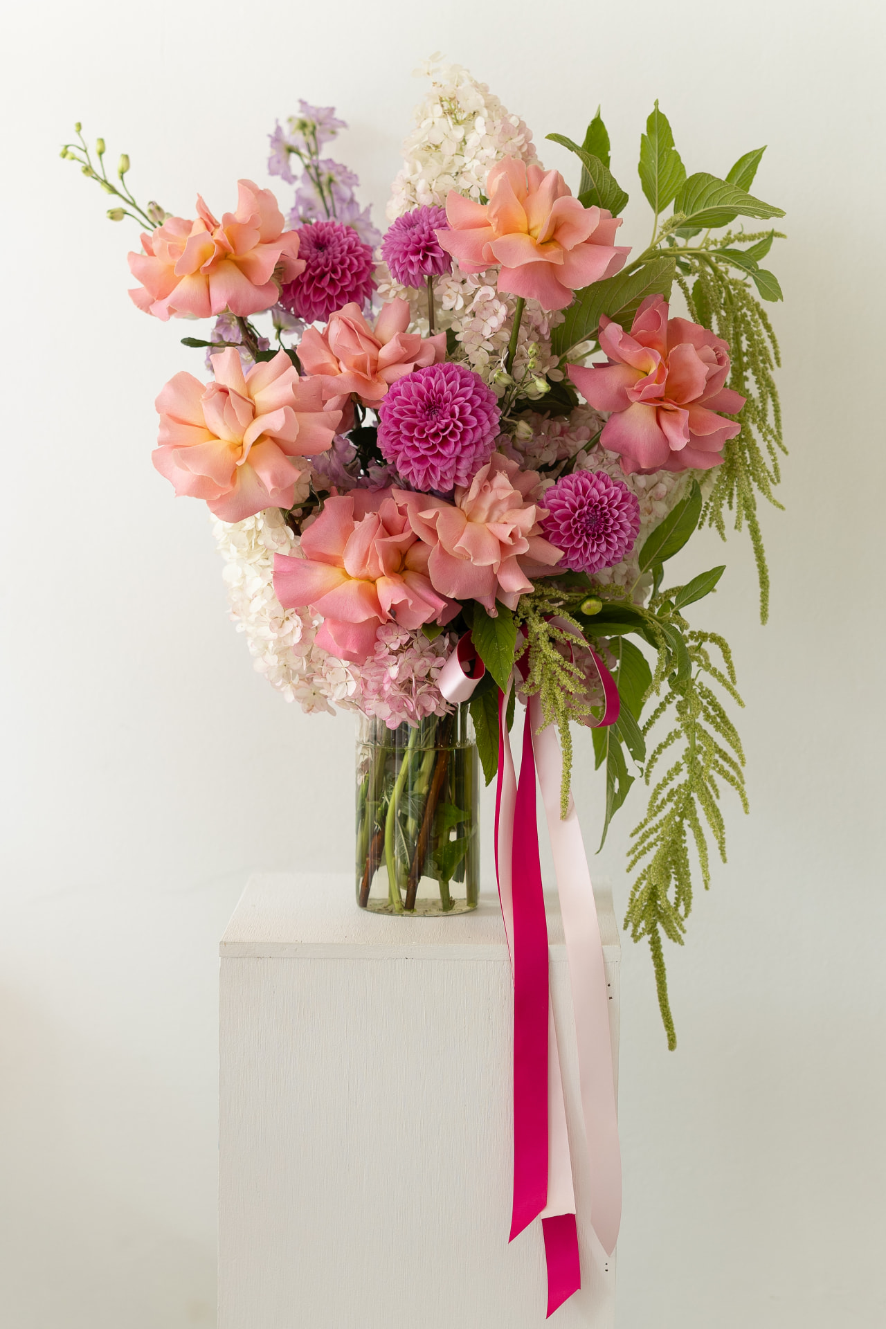 Tall vase arrangement of peach roses, dahlias and trailing amaranthus on a white plinth